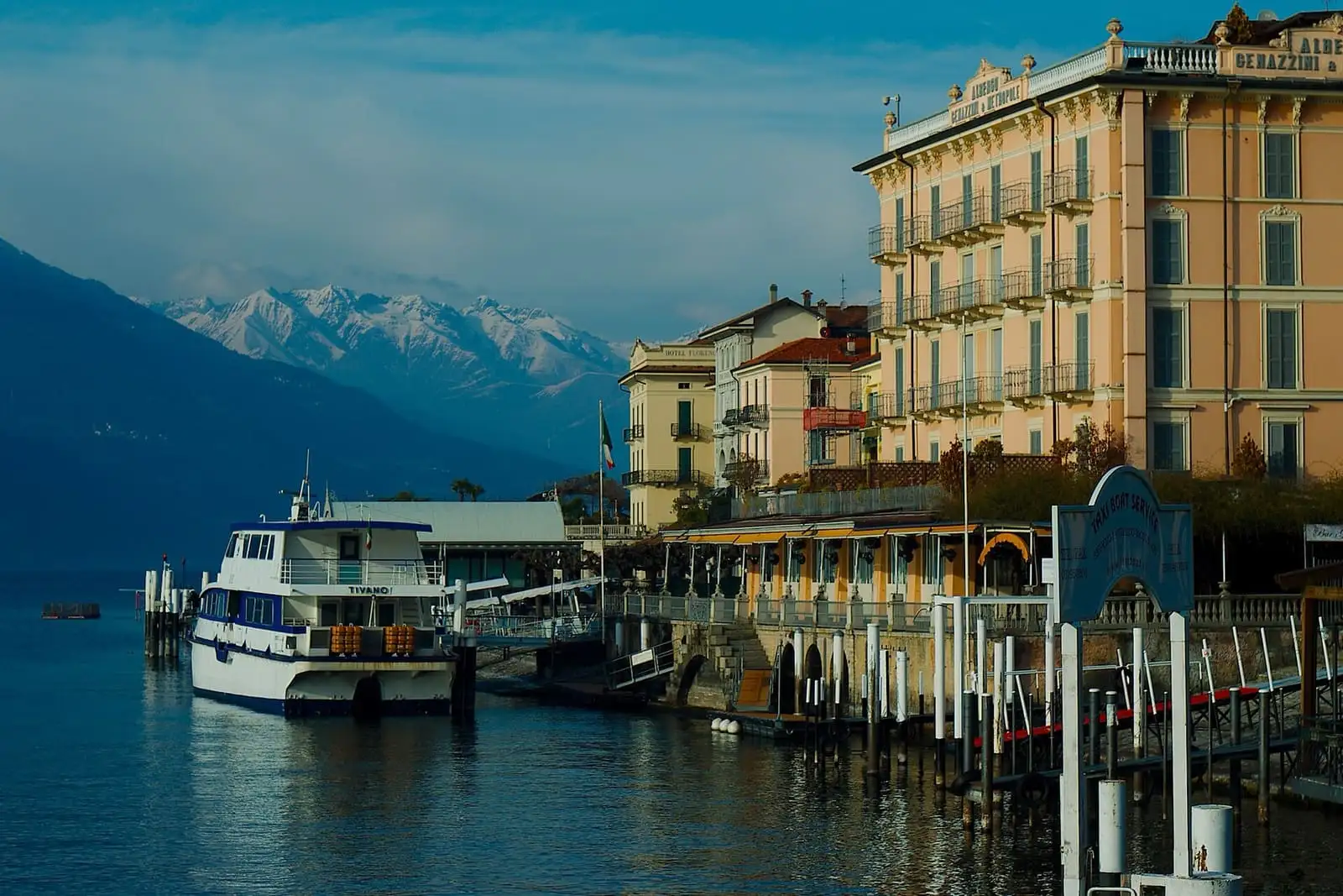 Lake Como ferry moored at the Bellagio pier beside lakefront hotels, with snowy peaks behind