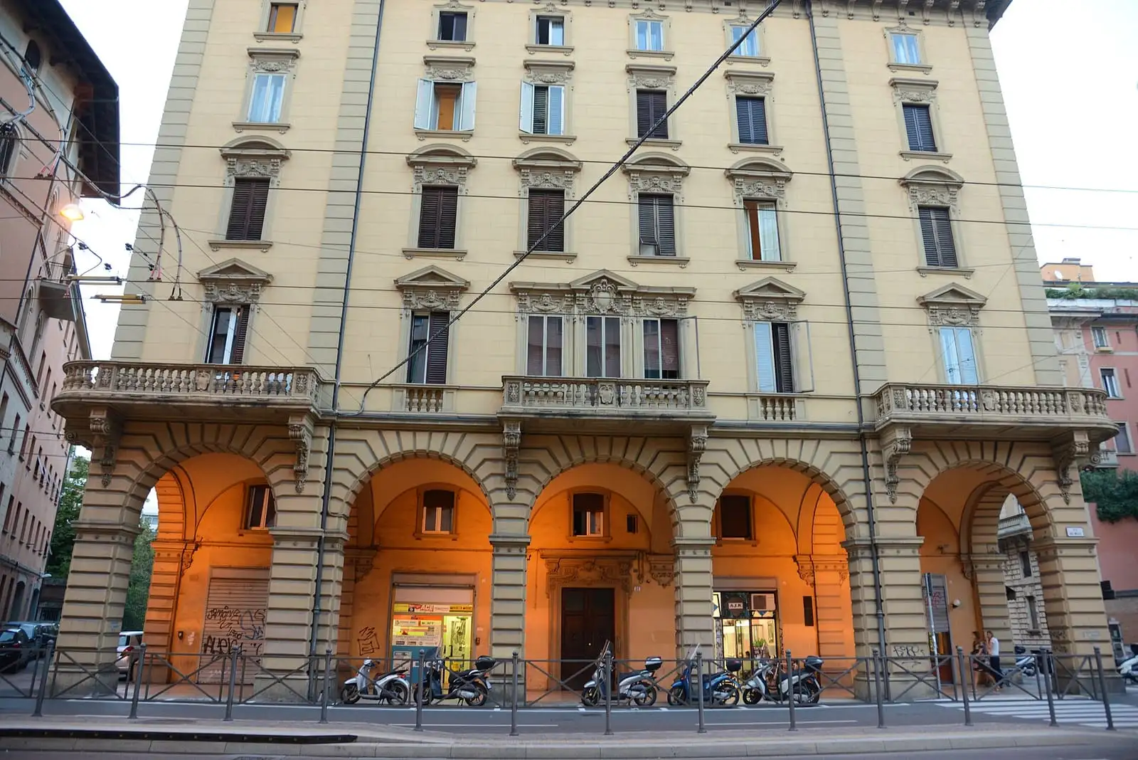 A row of lit arched porticoes under a yellow apartment building on Via dei Mille in Bologna