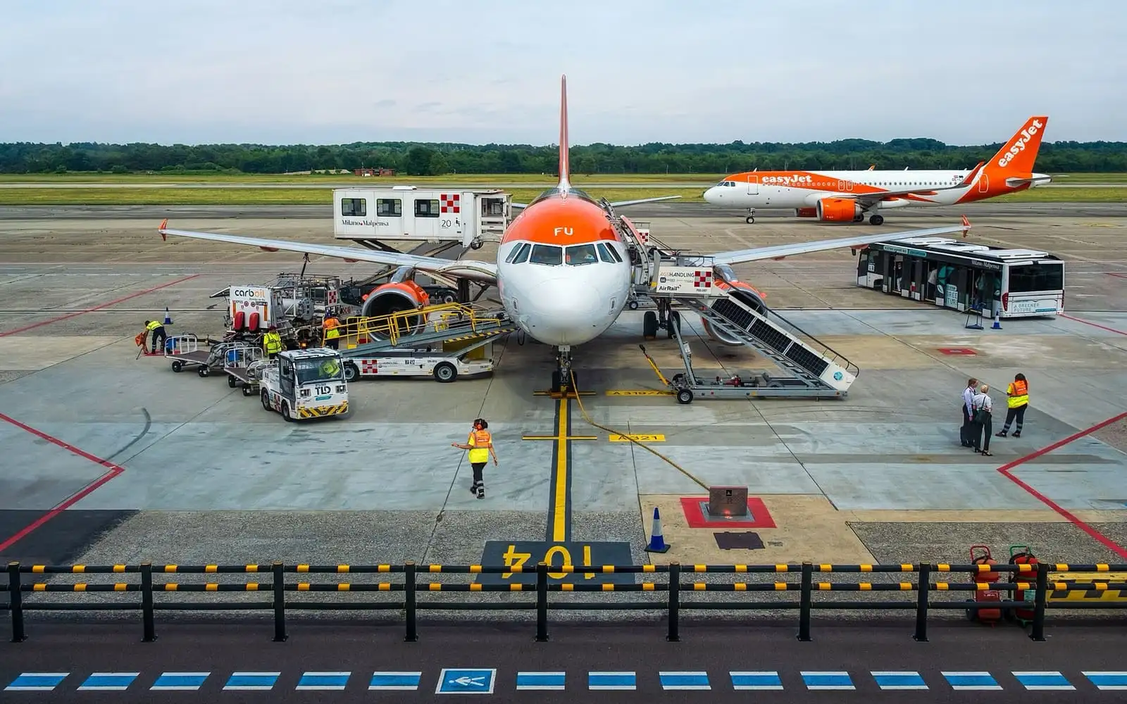 easyJet Airbus parked at a Malpensa Terminal 2 stand with ground equipment, a shuttle bus and a second easyJet jet taxiing behind