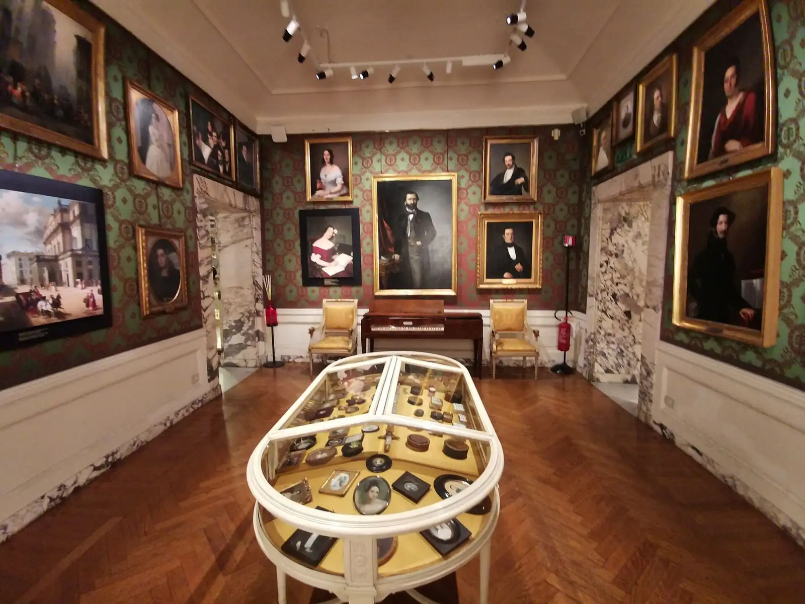 A room of the Museo Teatrale alla Scala with framed portraits on green walls, a piano and a glass display case in the center