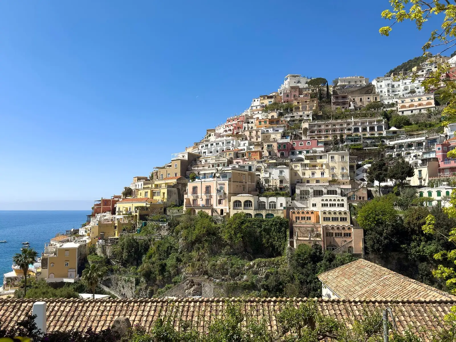 Pastel houses of Positano stacked up a green hillside above the sea, with tiled roofs in the foreground