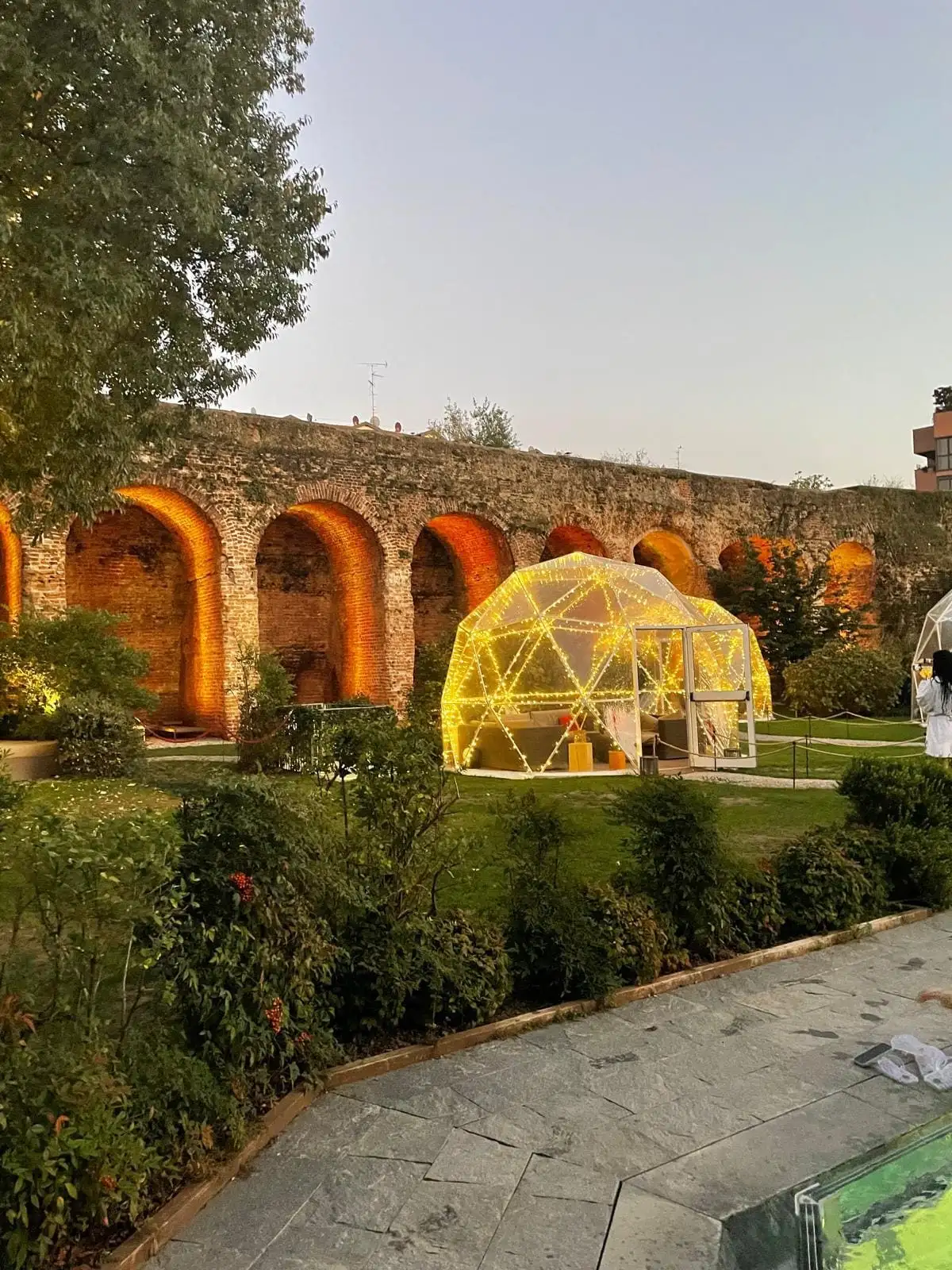 Il giardino di QC Spa Milano al tramonto, con una cupola illuminata davanti agli archi delle mura spagnole