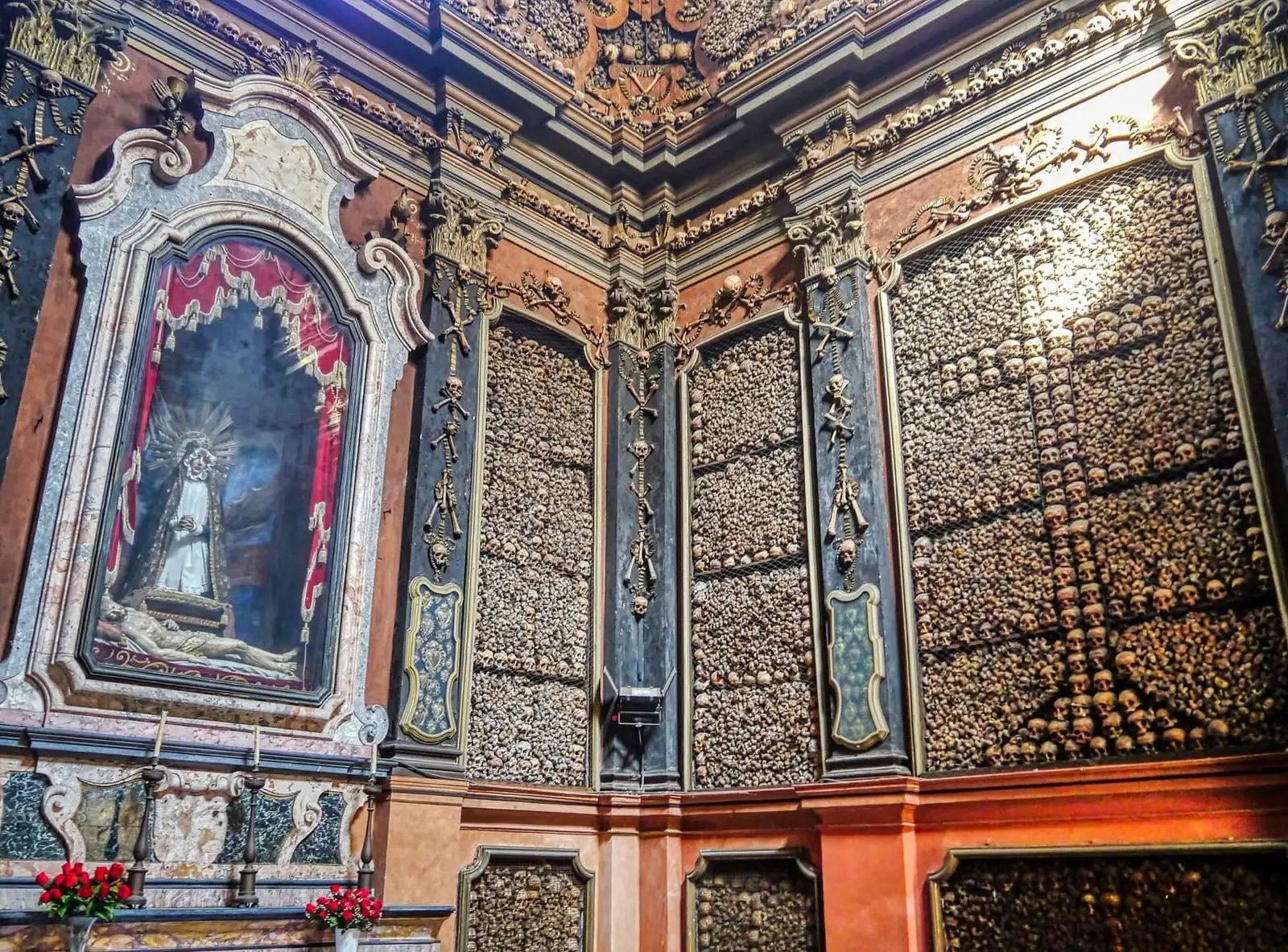 Walls of the ossuary chapel of San Bernardino alle Ossa covered in skulls and bones, with a framed statue of the Virgin on the left