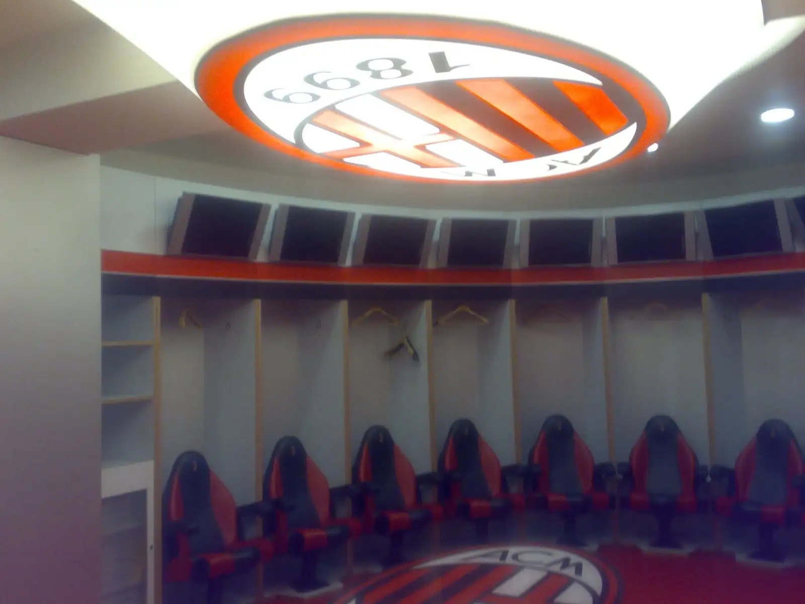 The AC Milan dressing room at San Siro in 2008, with red and black seats along the wall and the club crest on the ceiling