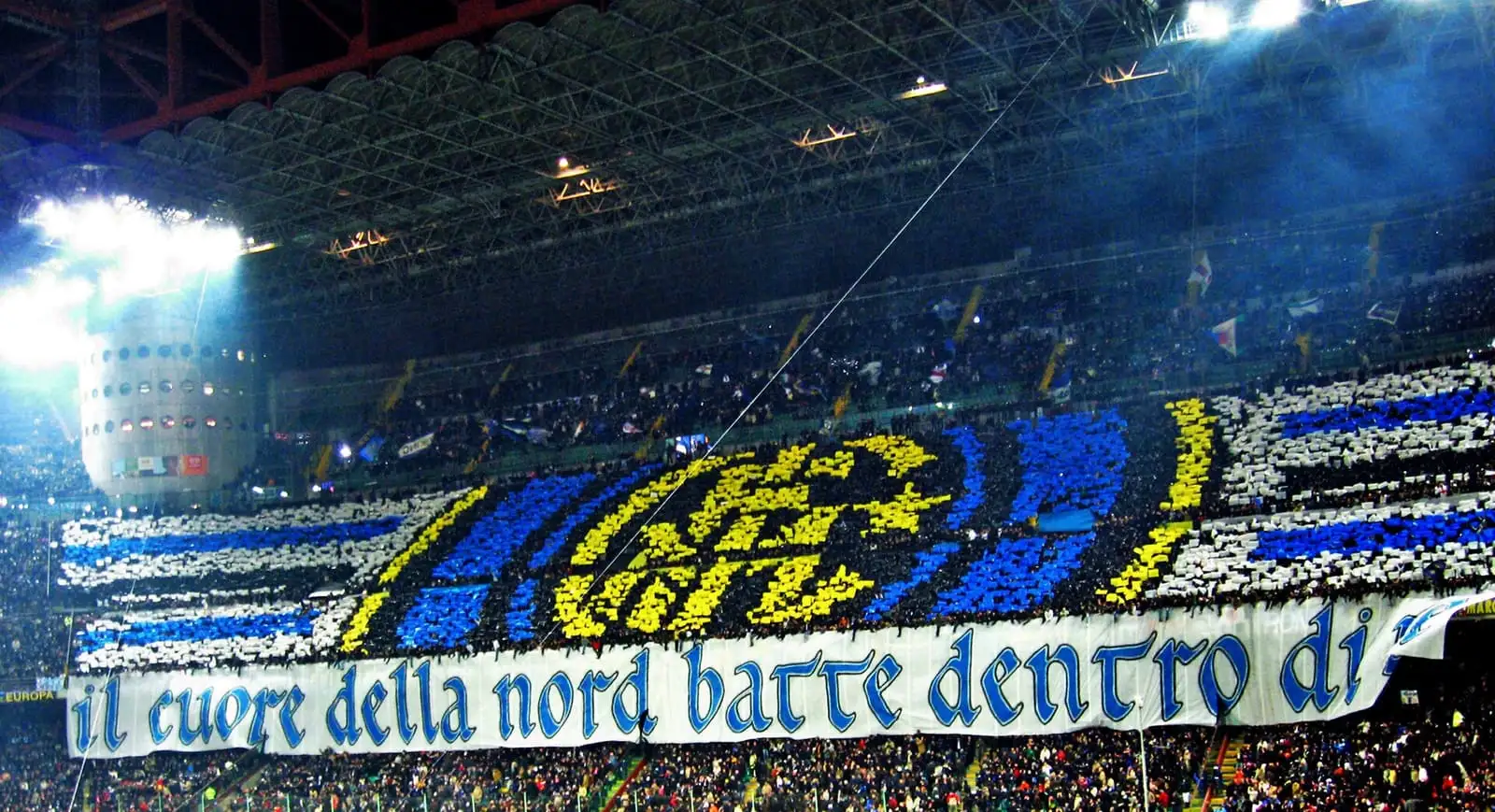 Inter fans in the Curva Nord at San Siro holding up a blue, white and gold card display of the club crest under the floodlights