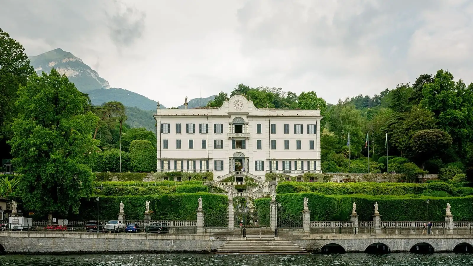 White facade of Villa Carlotta above its terraced garden stairs and the lakeside road, with mountains behind